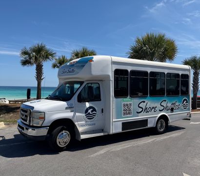 White shuttle bus parked at a sunny beachfront with palm trees, turquoise ocean and a clear blue sky.