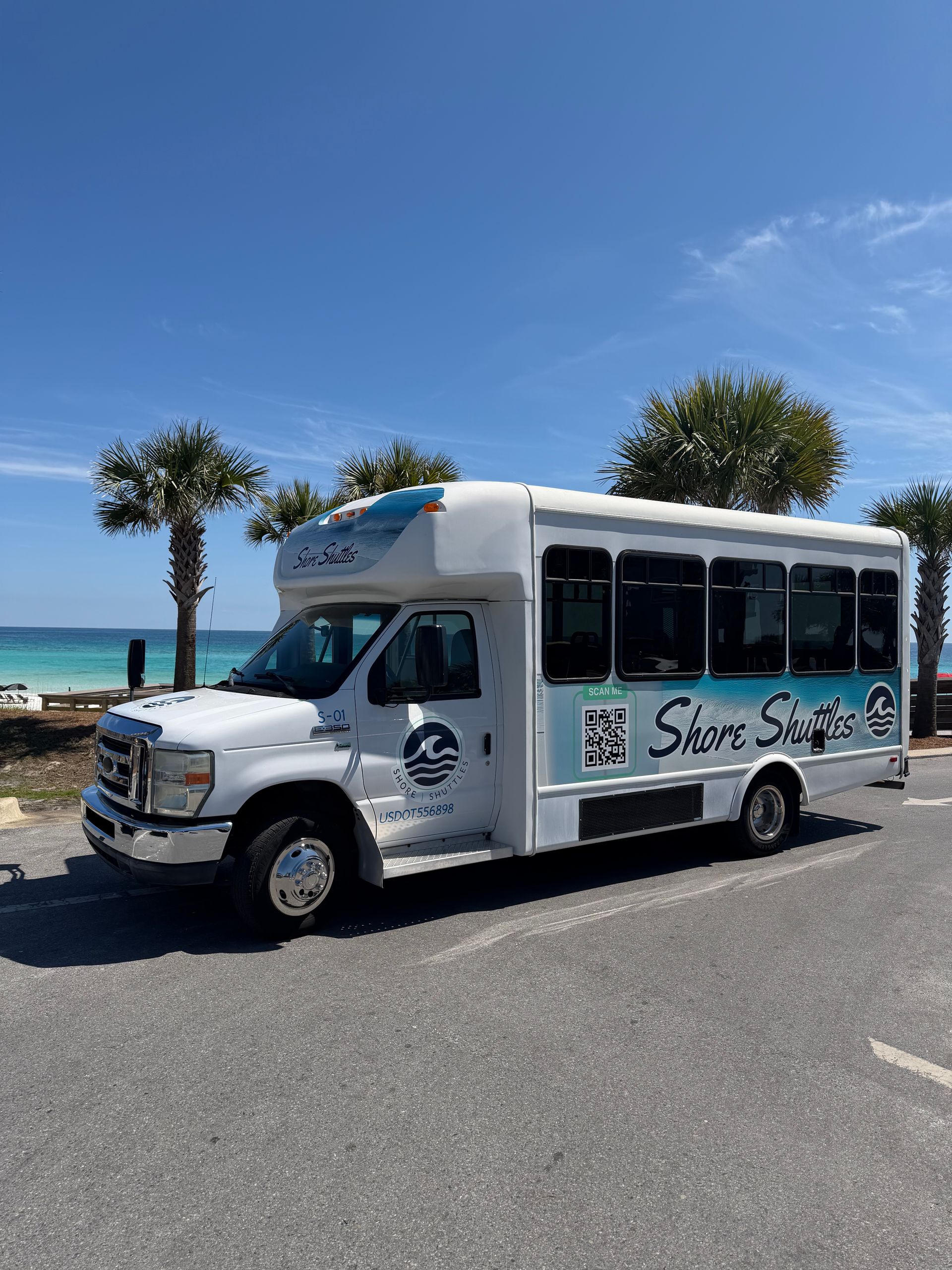 White shuttle bus parked at a sunny beachfront with palm trees, turquoise ocean and a clear blue sky.