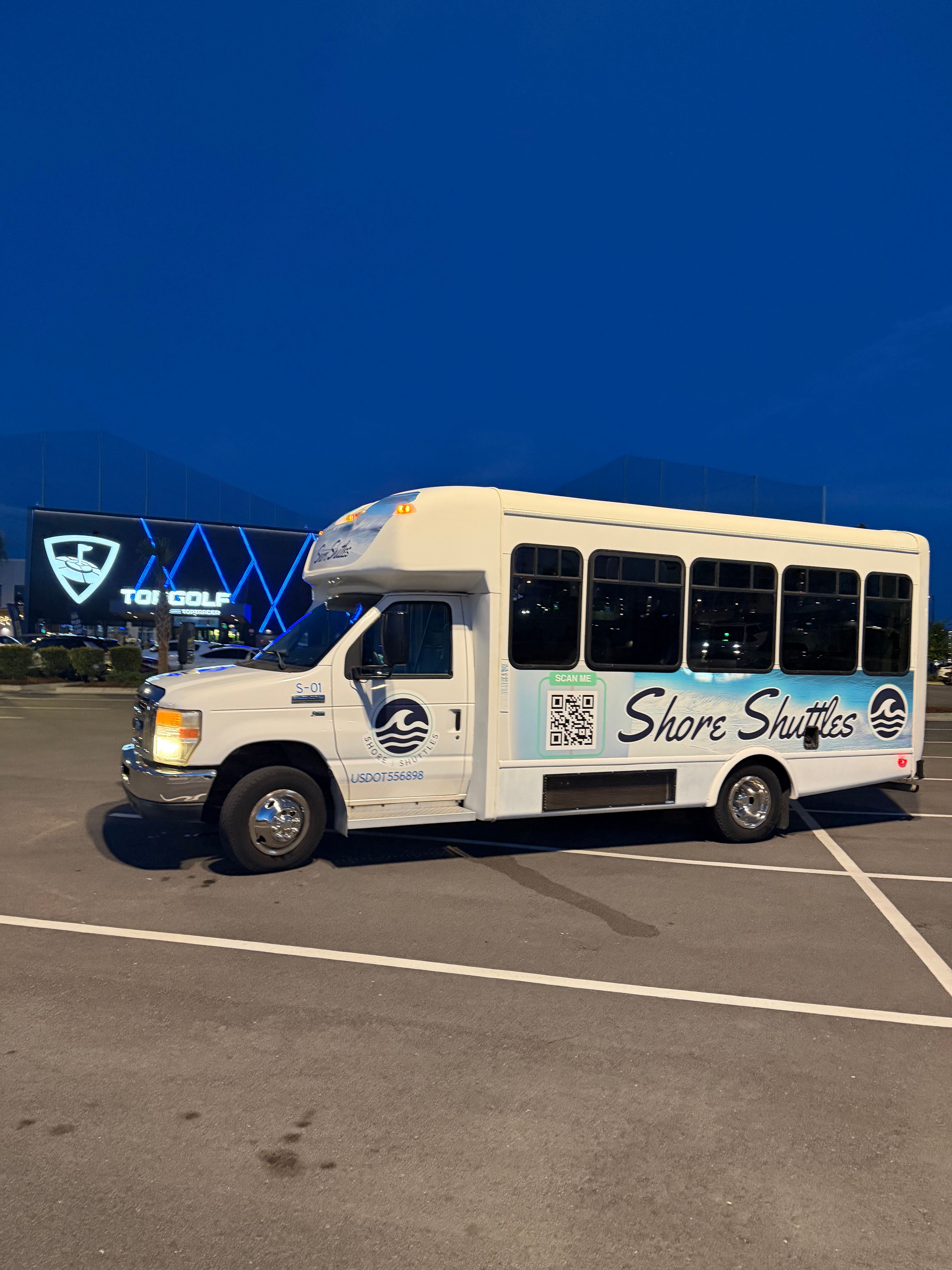 White passenger shuttle with blue wave logo and QR code parked in a lit parking lot at dusk, large entertainment building with blue neon geometric lights in the background.
