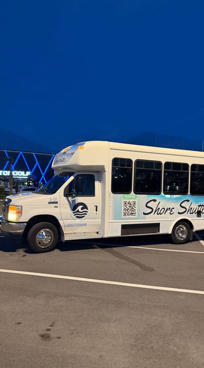 White passenger shuttle with blue wave logo and QR code parked in a lit parking lot at dusk, large entertainment building with blue neon geometric lights in the background.