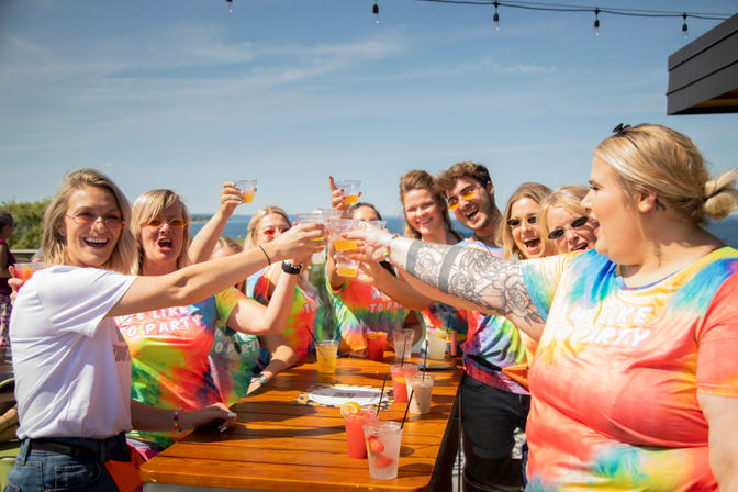 Cheerful group wearing colorful tie-dye shirts toasting with plastic cups on a sunny oceanfront rooftop patio, festive drinks on a wooden table.