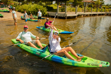 Two smiling adults in a bright green tandem kayak, woman reaching up playfully while paddling near a wooden dock on a sunny lake with other kayakers and shoreline greenery in the background.