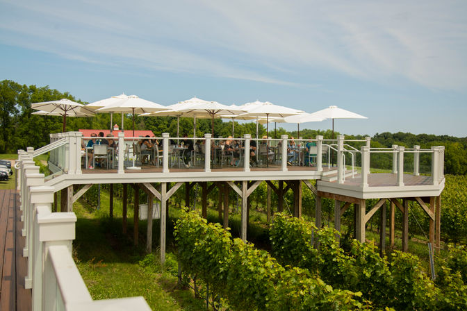 Elevated outdoor vineyard tasting deck with white umbrellas and diners seated behind glass railings, overlooking rows of grapevines and green countryside under a blue summer sky.