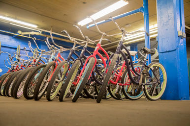 Colorful cruiser bicycles with wide tires and swept-back handlebars lined up indoors against blue pillars in a bike storage or rental area