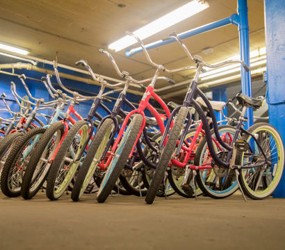 Colorful cruiser bicycles with wide tires and swept-back handlebars lined up indoors against blue pillars in a bike storage or rental area