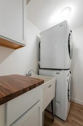 Bright compact home laundry room with stacked front-loading washer and dryer, white utility sink with chrome faucet, walnut butcher-block countertop and white cabinets