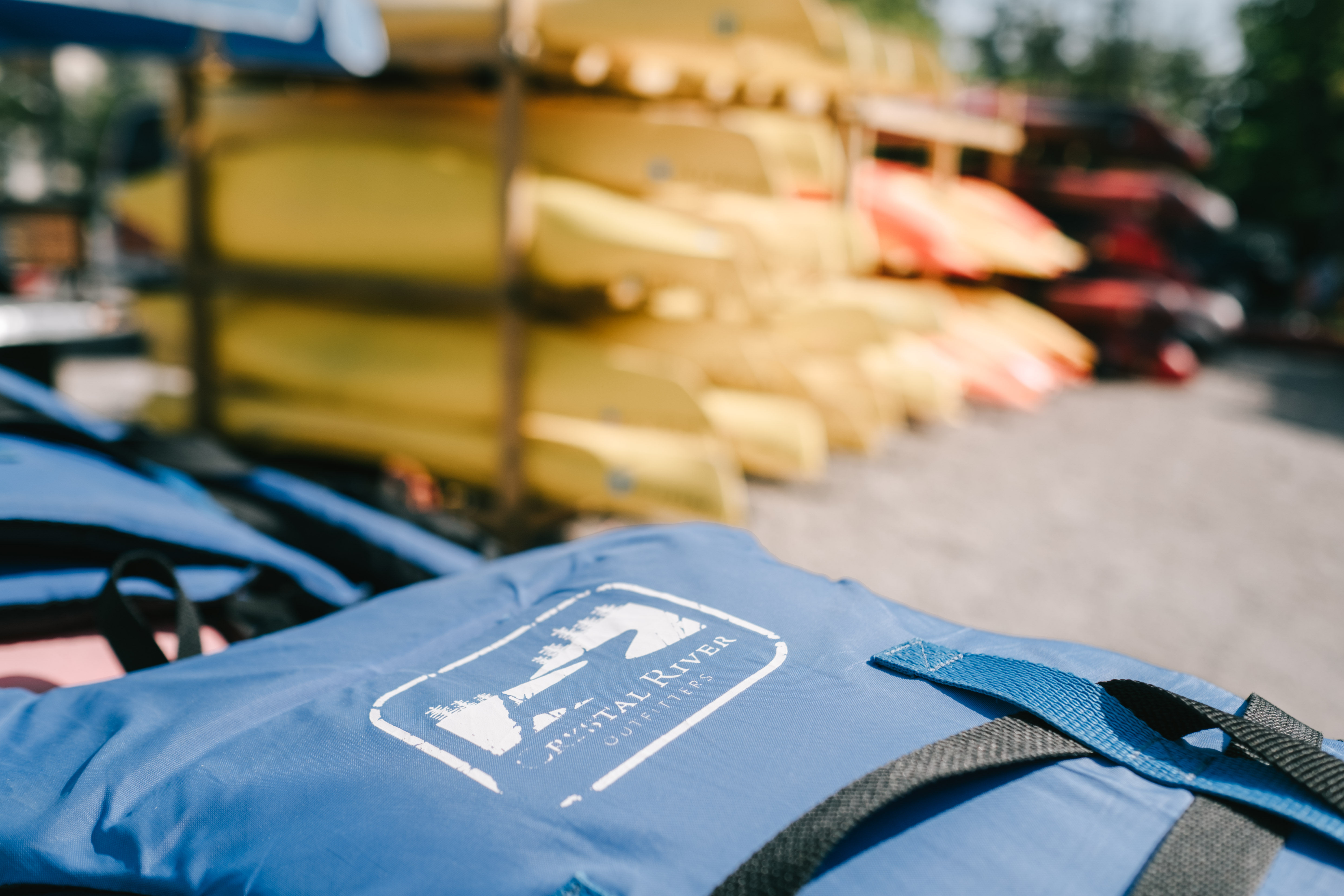 Close-up of a bright blue life jacket on a dock with stacked yellow and red kayaks blurred in the sunny background, outdoor watersports rental scene