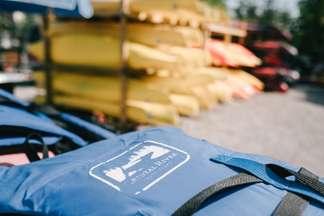Close-up of a bright blue life jacket on a dock with stacked yellow and red kayaks blurred in the sunny background, outdoor watersports rental scene