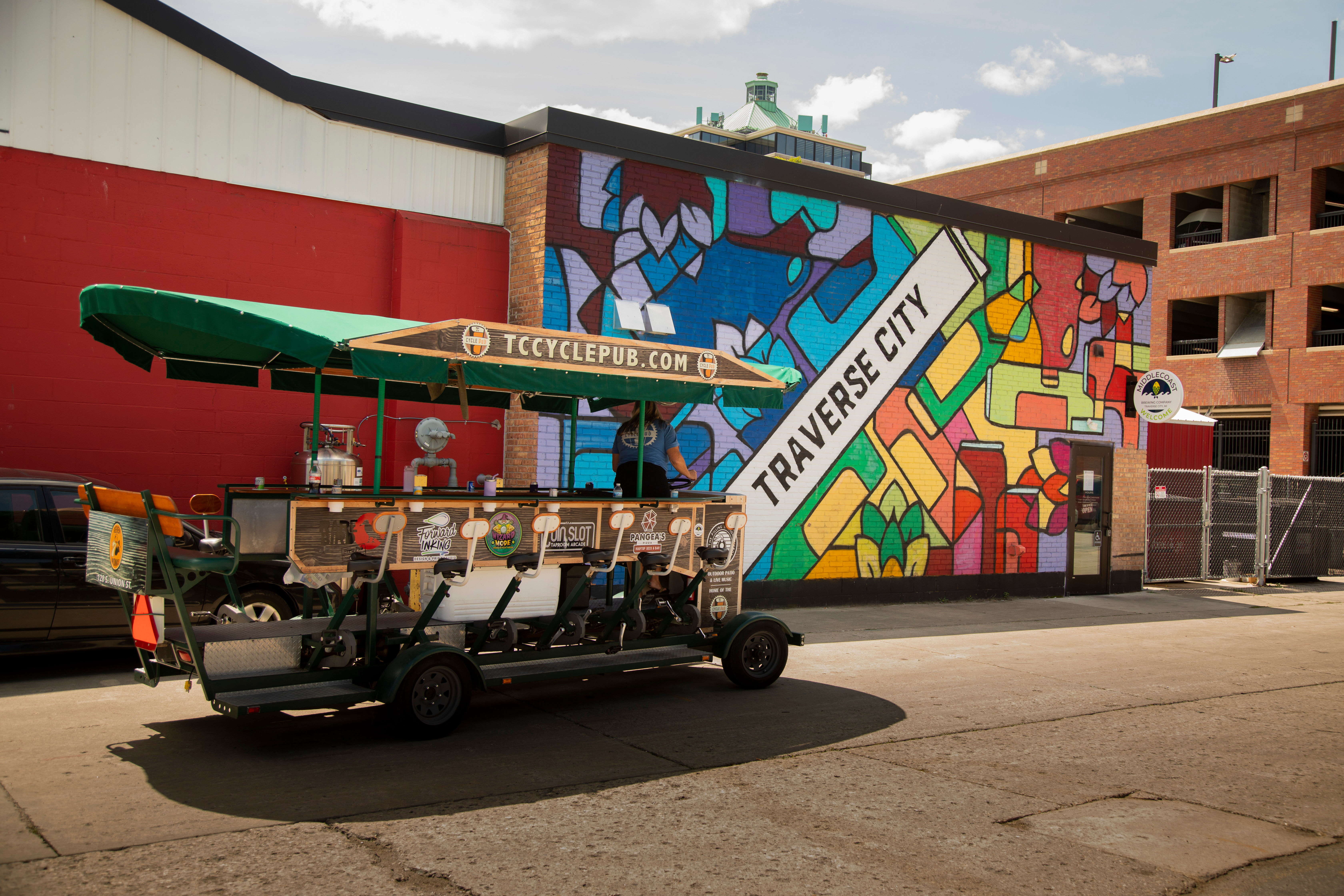 Colorful geometric mural reading Traverse City on a brick wall beside a green-canopied pedal pub bike parked on a sunny downtown street.