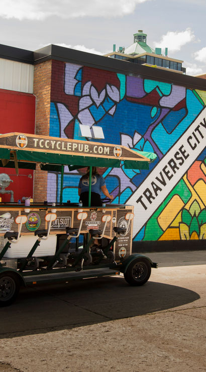Colorful geometric mural reading Traverse City on a brick wall beside a green-canopied pedal pub bike parked on a sunny downtown street.