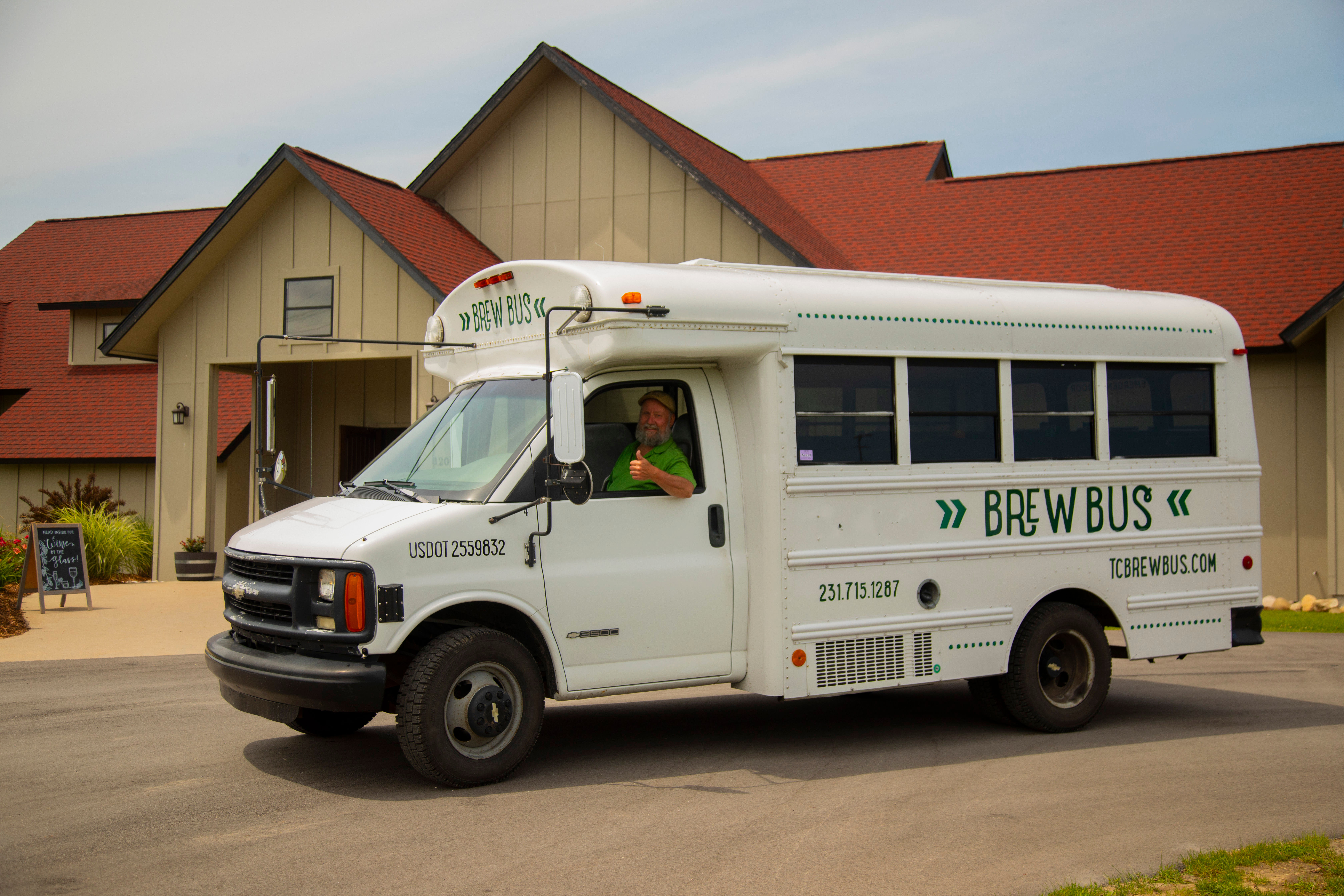 White brew-themed shuttle bus parked in front of a tan building with red roofs, driver giving a thumbs-up from the open window.