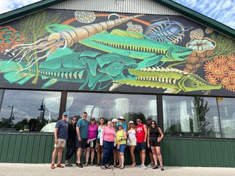 Bright coastal street-art mural of oversized green fish and sea creatures painted on a building facade above a storefront; a smiling group of adults in summer clothes poses in front.