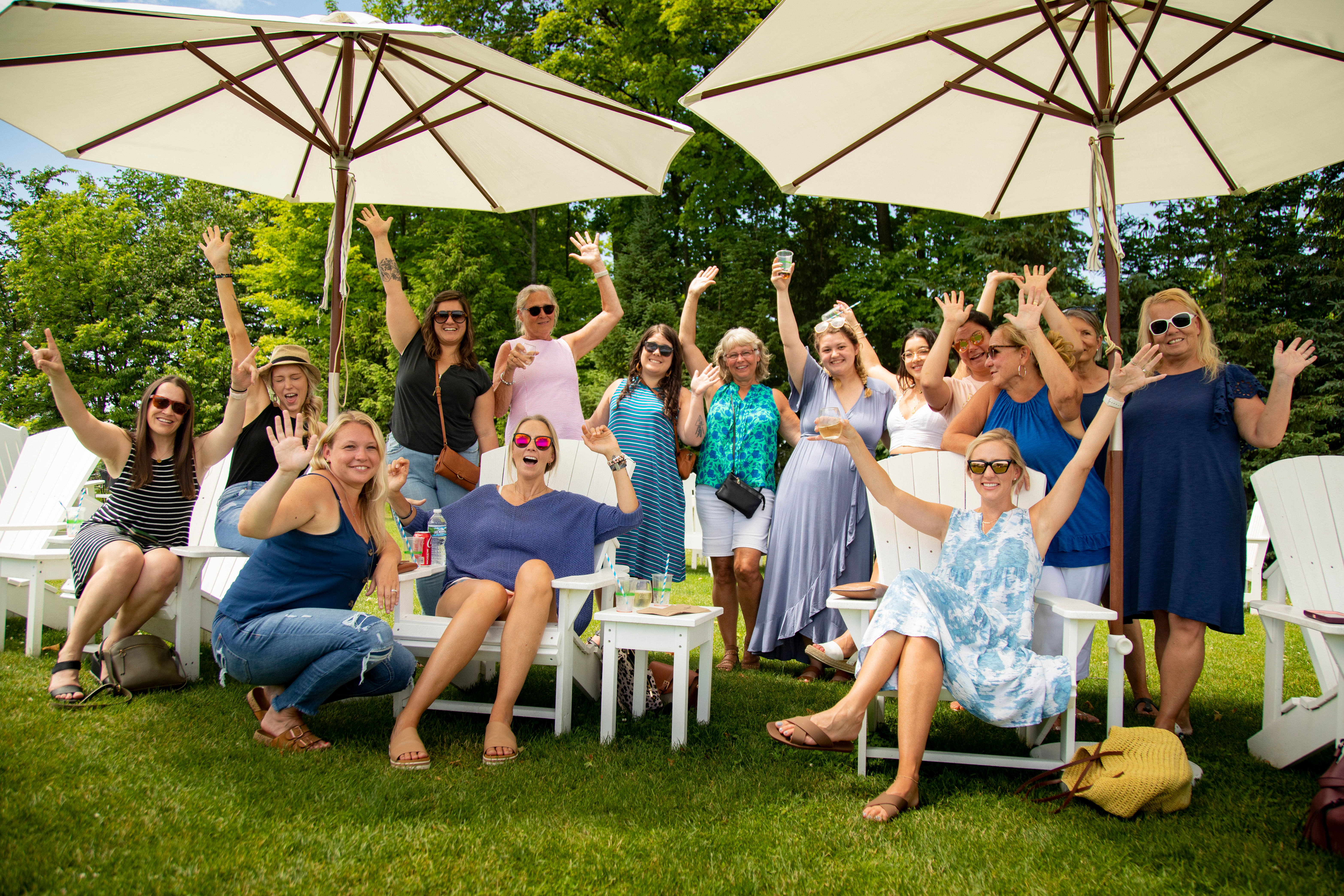 Group of smiling women raising their hands at an outdoor summer lawn party, seated in white Adirondack chairs under large patio umbrellas.