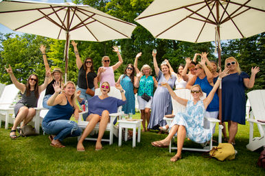 Group of smiling women raising their hands at an outdoor summer lawn party, seated in white Adirondack chairs under large patio umbrellas.