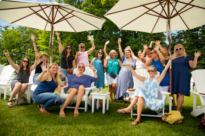 Group of smiling women raising their hands at an outdoor summer lawn party, seated in white Adirondack chairs under large patio umbrellas.
