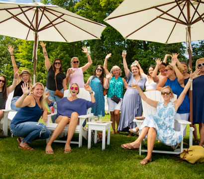 Group of smiling women raising their hands at an outdoor summer lawn party, seated in white Adirondack chairs under large patio umbrellas.