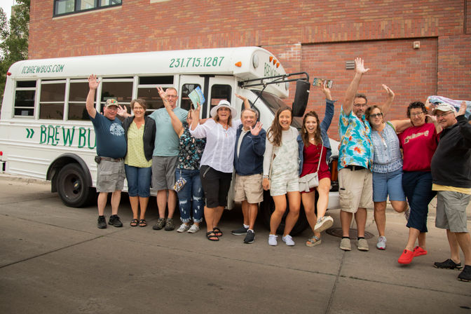 Cheerful mixed-age group in casual summer clothes smiling and waving while posing in front of a white tour bus parked on a city street beside a brick building.