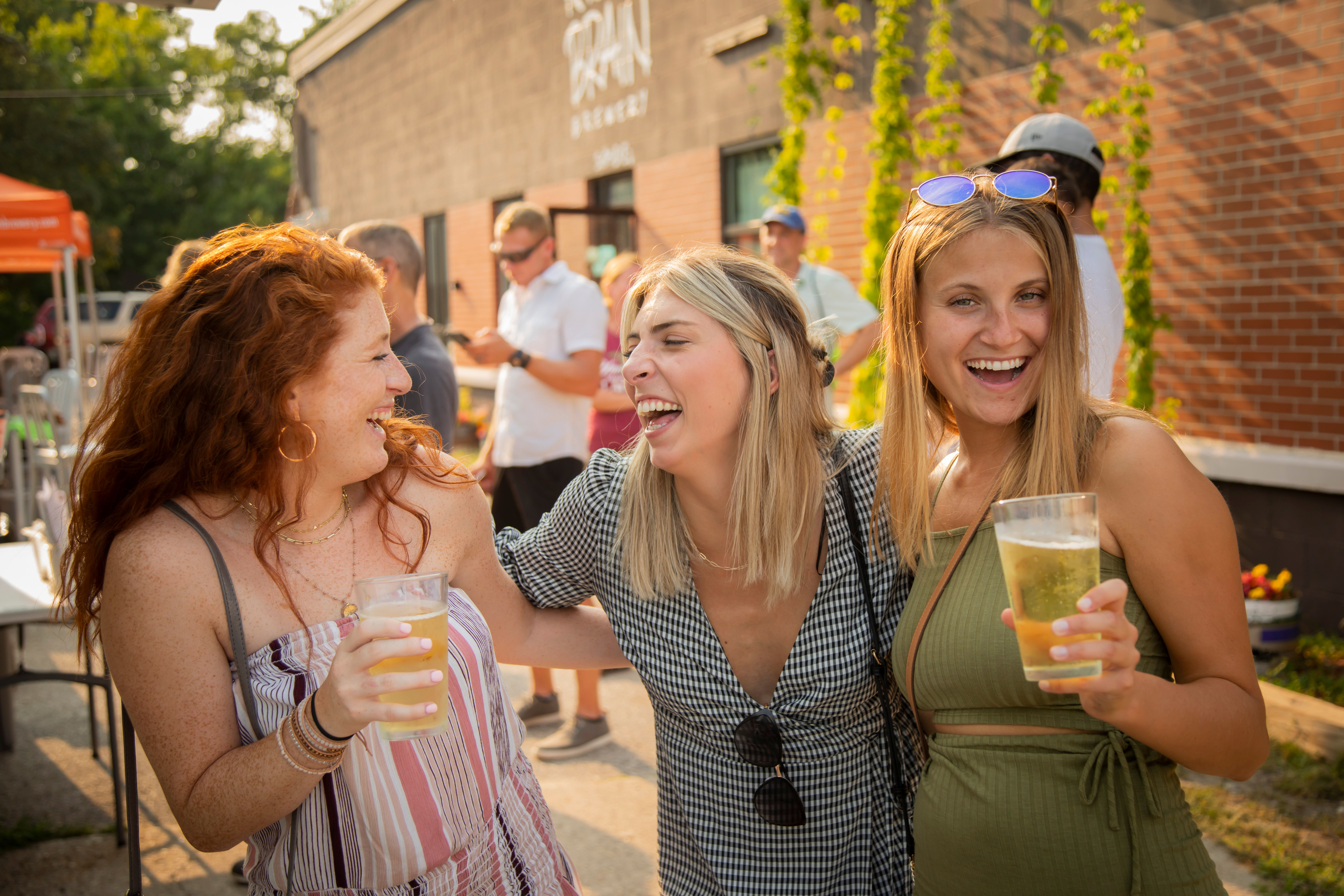 Three young women laughing together on a sunny outdoor patio, each holding a plastic cup of beer at a summer beer garden or street festival with a brick building backdrop.