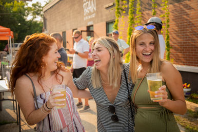 Three young women laughing together on a sunny outdoor patio, each holding a plastic cup of beer at a summer beer garden or street festival with a brick building backdrop.