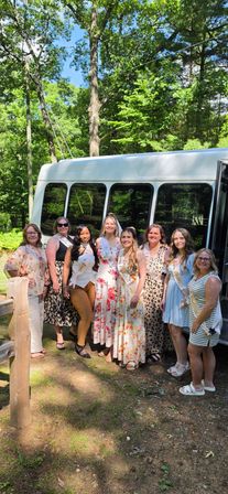 Eight women in summer dresses and sashes posing in front of a white shuttle bus in a sunlit wooded park — outdoor bachelorette party group photo.