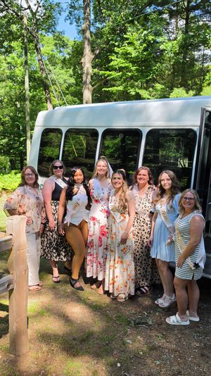 Eight women in summer dresses and sashes posing in front of a white shuttle bus in a sunlit wooded park — outdoor bachelorette party group photo.