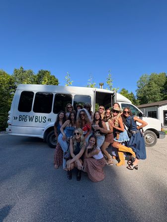 Smiling group of women posing in front of a white "Brew Bus" shuttle on a sunny summer day in a tree-lined parking area — celebratory outing