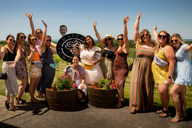 Cheerful group of women posing at a sunny vineyard outing, wearing colorful summer dresses and fanny packs, holding oversized face cutouts and a round sign, with wooden flower planters in the foreground and rows of grapevines under a blue sky.