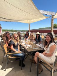 Group of friends at an outdoor winery patio tasting, women seated around a wooden table with wine flights under beige shade sails and a clear blue sky with countryside beyond.