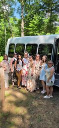Bachelorette party group photo — bride in a veil and sash with friends in floral and patterned summer dresses and sashes, smiling by a white party shuttle bus in a sunlit wooded park.