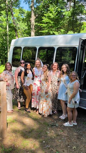Bachelorette party group photo — bride in a veil and sash with friends in floral and patterned summer dresses and sashes, smiling by a white party shuttle bus in a sunlit wooded park.