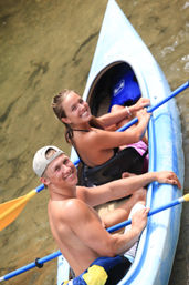 Close-up overhead view of two smiling young adults kayaking in a blue tandem kayak on shallow river water, holding blue paddles on a sunny summer day