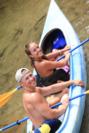Close-up overhead view of two smiling young adults kayaking in a blue tandem kayak on shallow river water, holding blue paddles on a sunny summer day