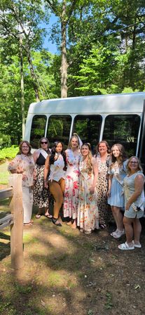 Outdoor bachelorette group photo: women in sashes and summer dresses, bride-to-be wearing a veil, posing in front of a white shuttle bus in a sunny wooded park.