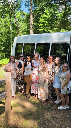 Outdoor bachelorette group photo: women in sashes and summer dresses, bride-to-be wearing a veil, posing in front of a white shuttle bus in a sunny wooded park.