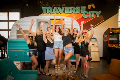 Group of seven women in sunglasses cheering with raised arms in front of a turquoise vintage camper inside a Traverse City indoor venue, casual summer outfits, one wearing a sash, playful celebration under colorful mural signage.