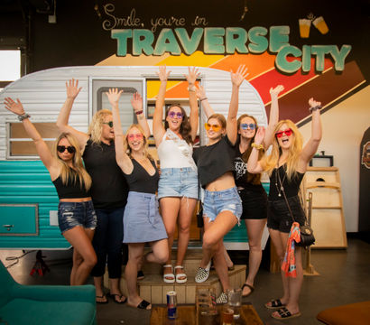 Group of seven women in sunglasses cheering with raised arms in front of a turquoise vintage camper inside a Traverse City indoor venue, casual summer outfits, one wearing a sash, playful celebration under colorful mural signage.