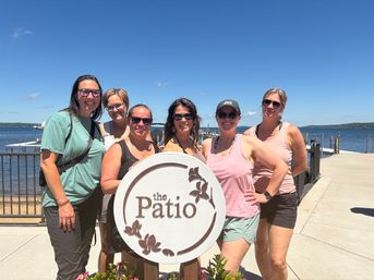 Six friends in sunglasses smiling by a round wooden patio sign on a sunny lakefront dock with calm water and clear blue sky
