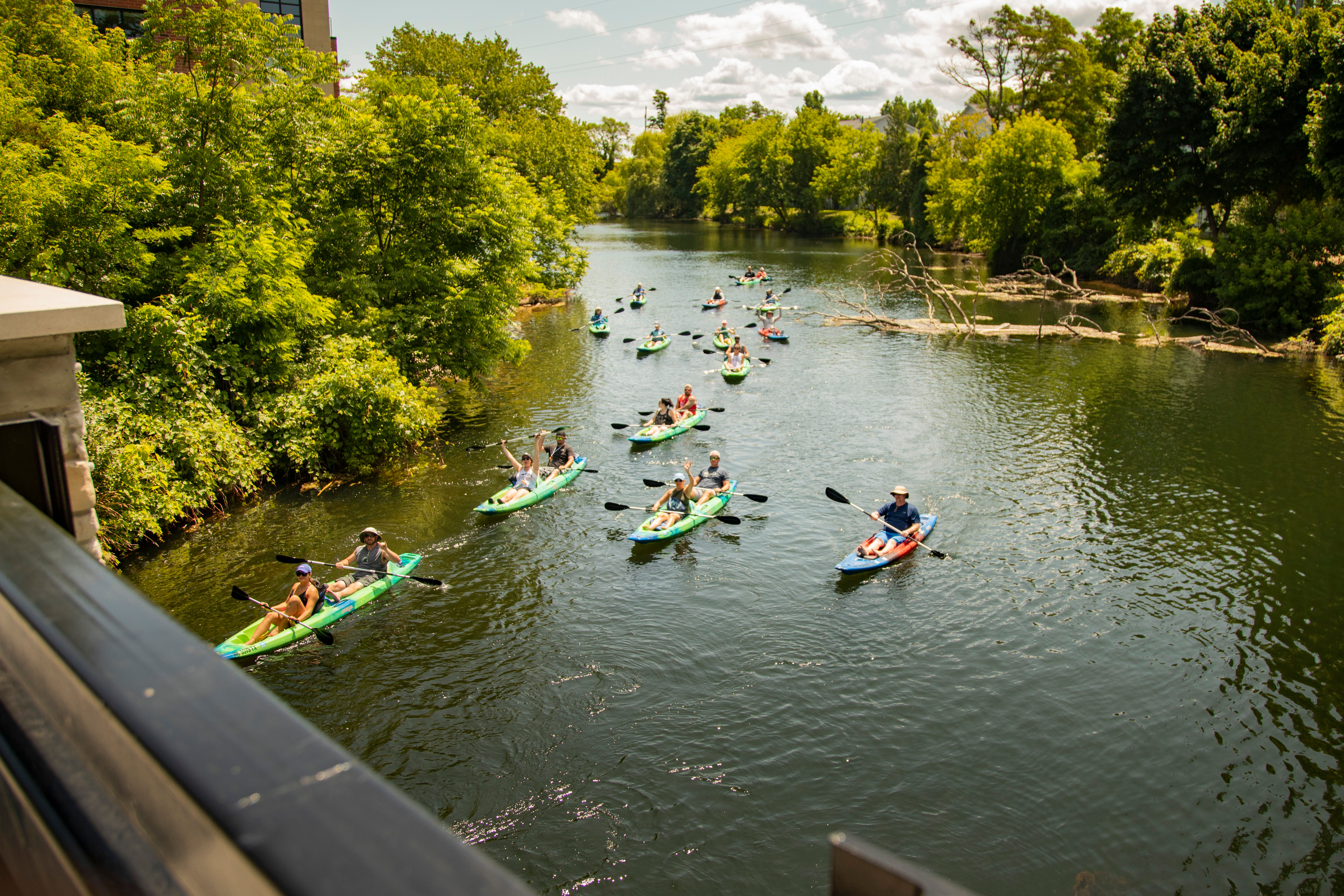 Group of kayakers paddling colorful single and tandem kayaks down a tree-lined river on a sunny summer day, viewed from a bridge.