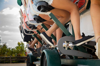 Close-up of several people’s legs and sneakers pedaling a multi-seat pedal-powered party bike on an outdoor group bike tour with trees and a wooden fence in the background