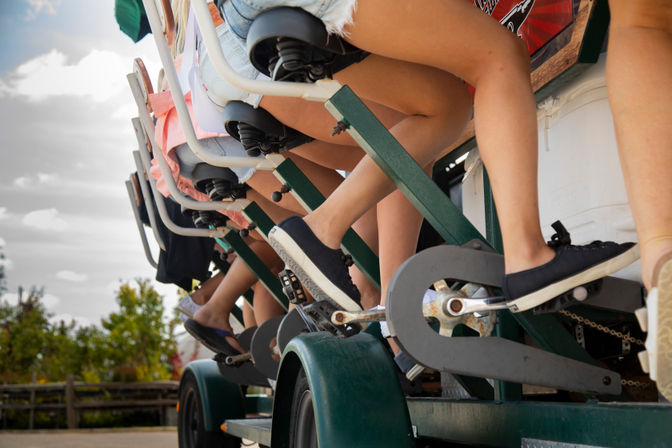Close-up of several people’s legs and sneakers pedaling a multi-seat pedal-powered party bike on an outdoor group bike tour with trees and a wooden fence in the background