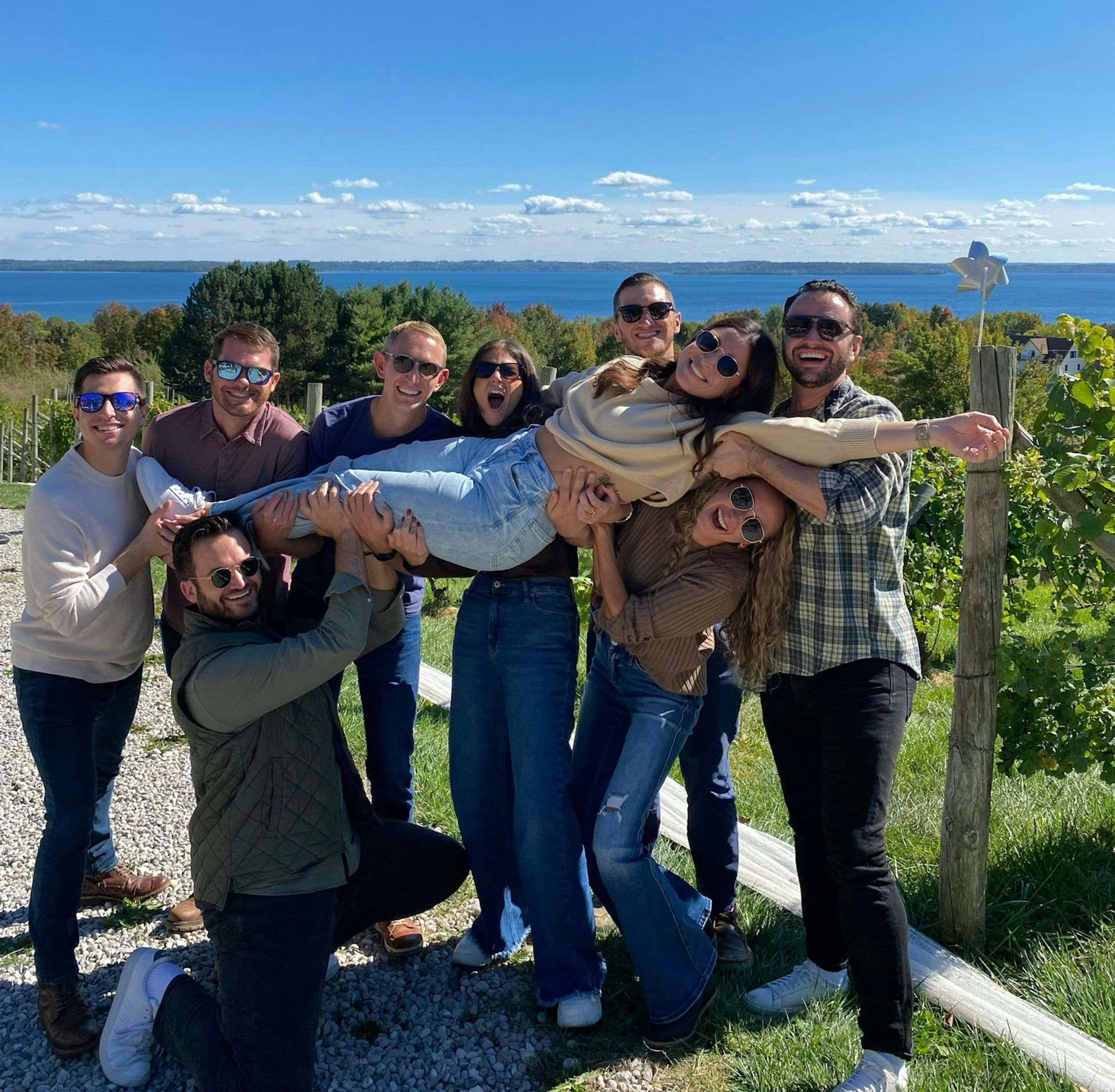 Group of friends laughing in a lakeside vineyard on a sunny fall day, holding a smiling woman horizontally with blue water, rolling vines, and a clear sky in the background.