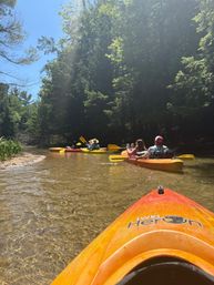 Sunny summer paddling on a shallow, clear forest river — orange kayak bow in the foreground, three colorful kayaks and paddlers drifting under a leafy green canopy.