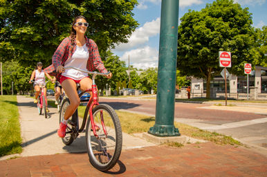 Three women riding red cruiser bicycles along a tree-lined city sidewalk on a sunny summer day, smiling in casual clothes and sunglasses