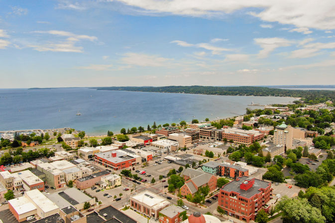 Aerial view of a lakefront downtown with marina and sailboats, brick commercial buildings, tree-lined streets and a shoreline under a blue partly-cloudy sky.