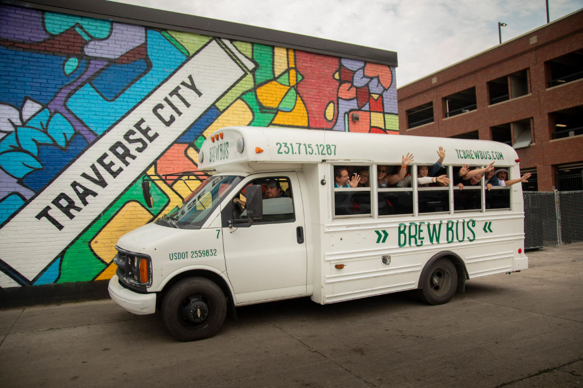 White shuttle-style tour bus with passengers waving from open windows parked beside a colorful Traverse City mural on an urban street.