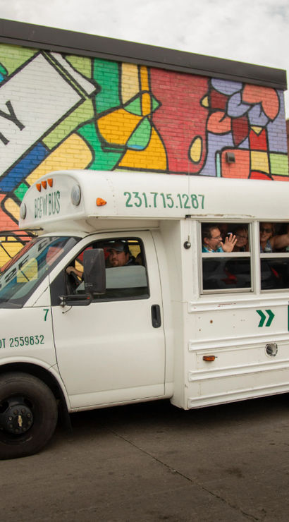 White shuttle-style tour bus with passengers waving from open windows parked beside a colorful Traverse City mural on an urban street.