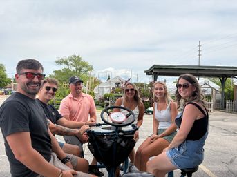 Group of six adults smiling and wearing sunglasses on a circular multi-seat pedal bike outside a garden center with greenhouses and potted plants on an overcast summer day.