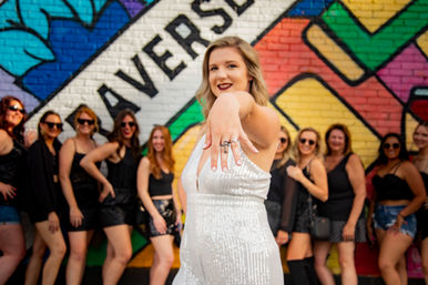 Woman in a white sequin outfit proudly shows her engagement ring to camera while a group of friends dressed in black pose in front of a colorful street mural — lively bachelorette/engagement celebration.