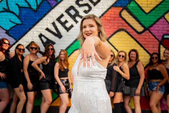 Woman in a white sequin outfit proudly shows her engagement ring to camera while a group of friends dressed in black pose in front of a colorful street mural — lively bachelorette/engagement celebration.