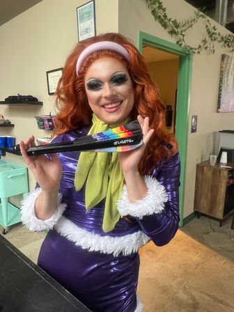 Smiling drag performer with red wig and dramatic makeup in a shiny purple dress with white trim and green scarf, holding a rainbow folding fan inside a bright indoor studio